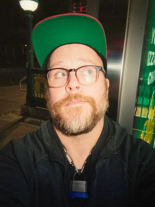 Selfie of me waiting for the bus in Queens, wearing a green-and-pink brimmed snapback cap flipped up high, glasses, and a ginger beard, looking up and to the side while standing outside at night near a streetlamp. A padlock hangs from my chain collar, peeking out from behind the pull of a black hoodie on my torso.