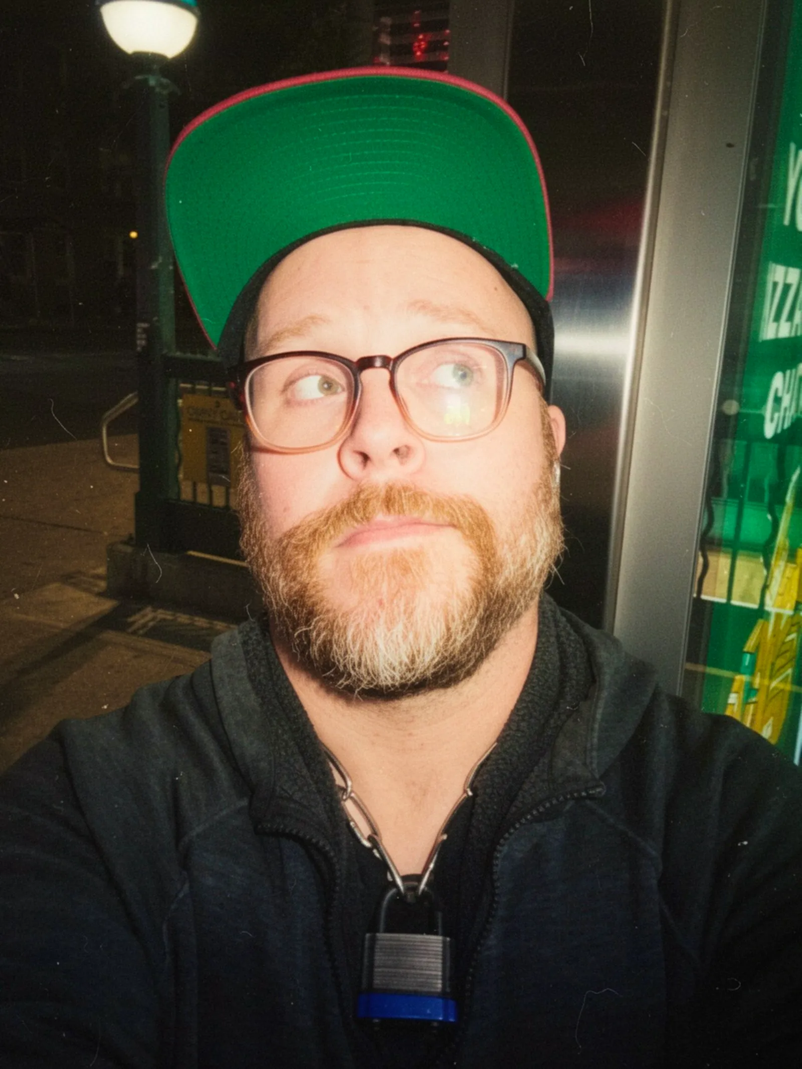 Selfie of me waiting for the bus in Queens, wearing a green-and-pink brimmed snapback cap flipped up high, glasses, and a ginger beard, looking up and to the side while standing outside at night near a streetlamp. A padlock hangs from my chain collar, peeking out from behind the pull of a black hoodie on my torso.