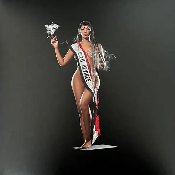 A woman stands barefoot on a small white platform against a dark charcoal background, wearing long beaded braids and a white pageant-style sash with red trim draped over her nude body, holding up a smoking object in one raised hand.