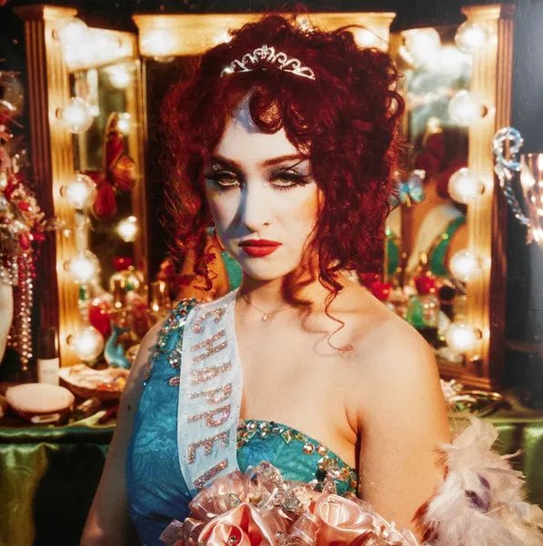 A red-haired woman in dramatic blue eye makeup wears a tiara, beaded sash and teal one-shoulder gown while holding a peach satin rose bouquet, posed before a warmly lit backstage vanity mirror ringed with glowing bulbs.