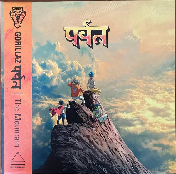 Four illustrated cartoon figures stand atop a steep rocky peak surrounded by vast pastel pink, blue and gold clouds, with Devanagari lettering above and a pink-orange vertical obi strip along the left edge.