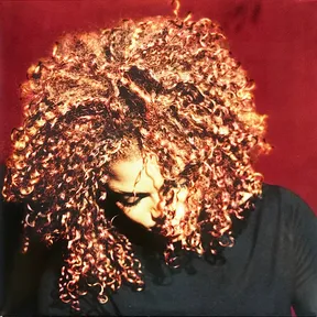 A woman with voluminous curly copper-toned hair tilts her head downward with eyes closed, her face partly veiled by ringlets, set against a deep crimson background and wearing a dark top.