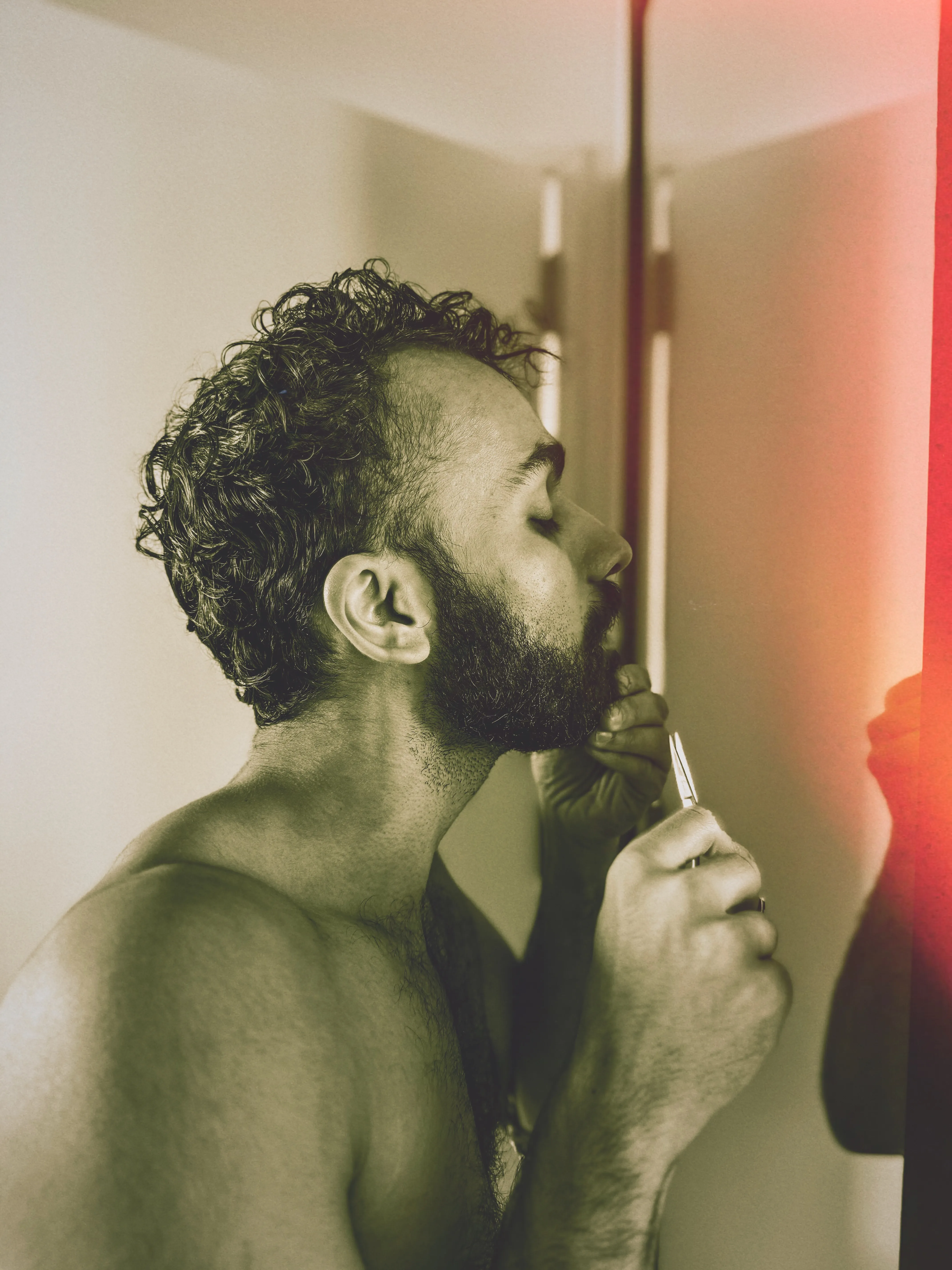 My partner, Sir Aditya, a lean and handsome brown-skinned Man with a a dark black and dense cropped beard stands shirtless in front of a mirror while primping and prepares to go out for the night by trimming His beard’s hairs with a pair of scissors.