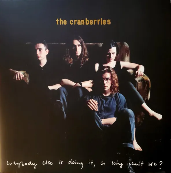 Four young people in dark clothing sit clustered on and around an armchair against a black background, dramatically lit in warm, moody tones, with one seated barefoot on the floor in front.