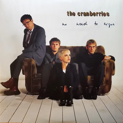 Four band members in dark suits pose on and around a worn brown velvet sofa in a stark white room with pale wooden floorboards, with handwritten-style text in the upper right corner.