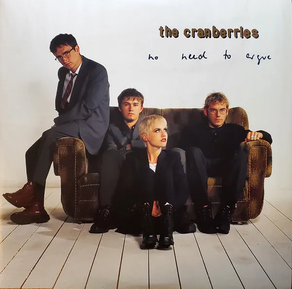 Four band members in dark suits pose on and around a worn brown velvet sofa in a stark white room with pale wooden floorboards, with handwritten-style text in the upper right corner.
