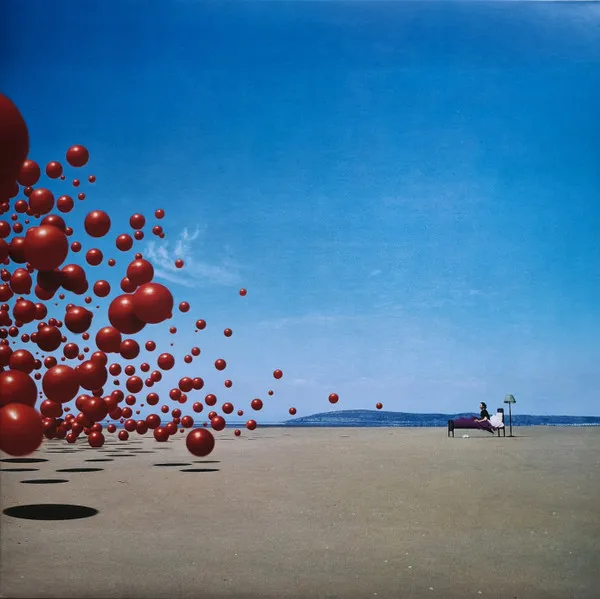 A surreal photographic scene of a vast pale beach under a deep blue sky, where a swarm of glossy red spheres tumbles in from the left casting dark round shadows, while a lone figure reclines on a bed beside a lamp at the right.