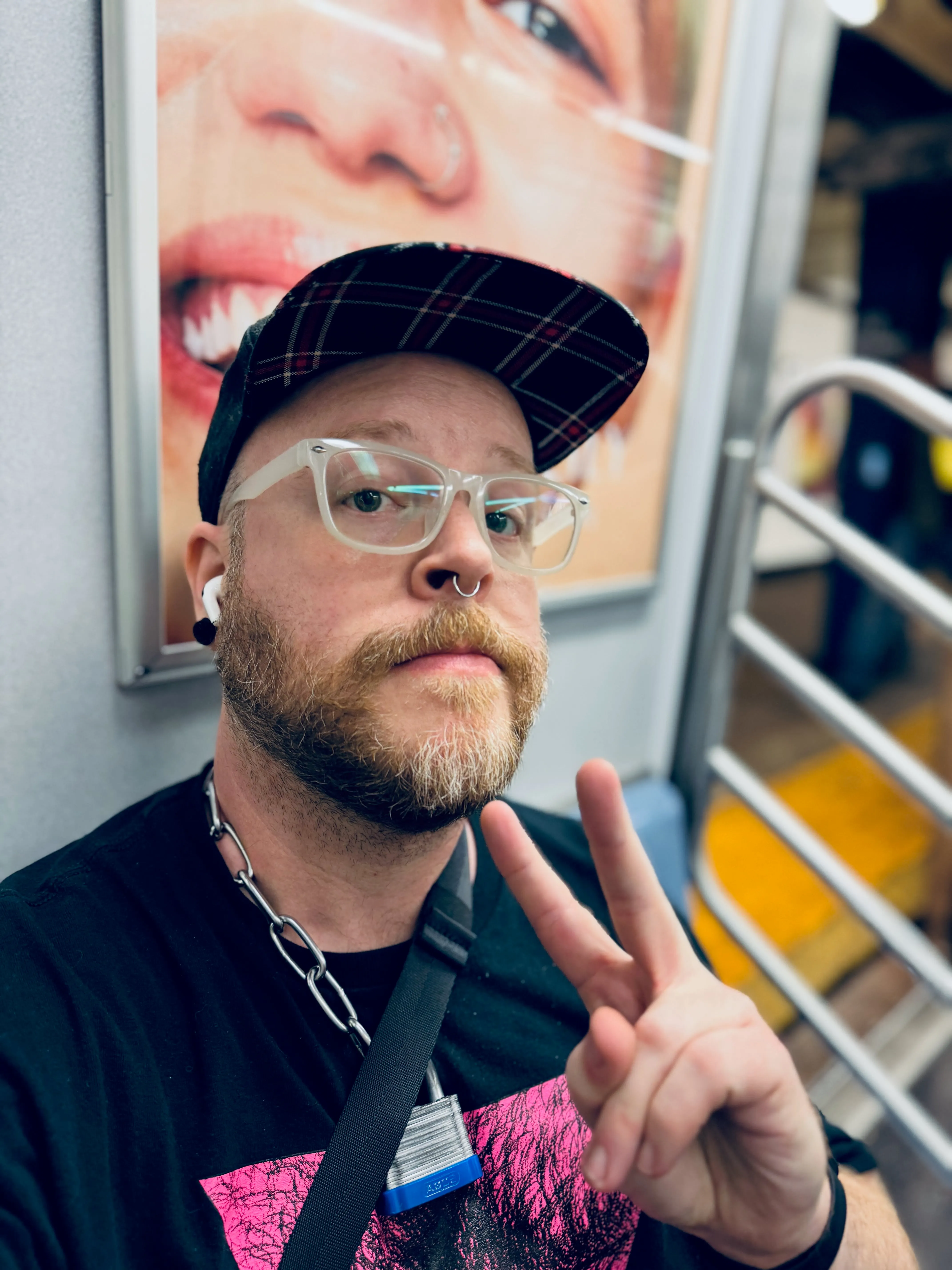 Selfie of me, a white masculine-presenting person with a red and white beard cropped tight flashing a peace sign. i am wearing a black snapback hat with a plaid brim, clear/white glasses, a septum ring, and a black t-shirt with a pink triangle showing a hairy illustrated chest. On top of that is my Sir’s chain and padlock collar around my neck. i’m flashing a peace sign to the camera as i sit in an open C train in New York.