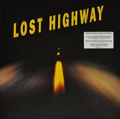 A dark, blurred nighttime photograph of a road stretching into blackness, with a glowing yellow-orange streak of light down the center and streaked headlight trails at the sides, overlaid with bold yellow block lettering at the top and a white sticker in the upper right corner.