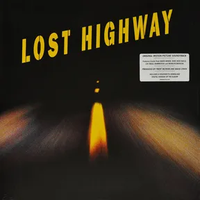 A dark, blurred nighttime photograph of a road stretching into blackness, with a glowing yellow-orange streak of light down the center and streaked headlight trails at the sides, overlaid with bold yellow block lettering at the top and a white sticker in the upper right corner.